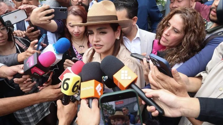 Una mujer con sombrero rodeada de periodistas y cámaras, hablando mientras sostenidos micrófonos de diferentes colores la rodean.