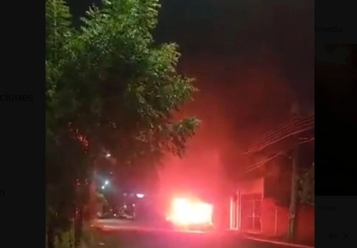 Un vehículo en llamas iluminando la calle por la noche, con árboles y edificios en el fondo.