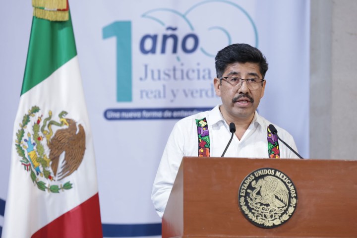 Un hombre con camiseta blanca y un chaleco colorido habla en un podio frente a una bandera mexicana y un fondo con el texto '1 año Justicia real y verde'.