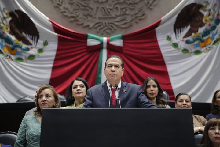 Un político hablando en un podio con una bandera mexicana detrás, rodeado de varias mujeres en la cámara.