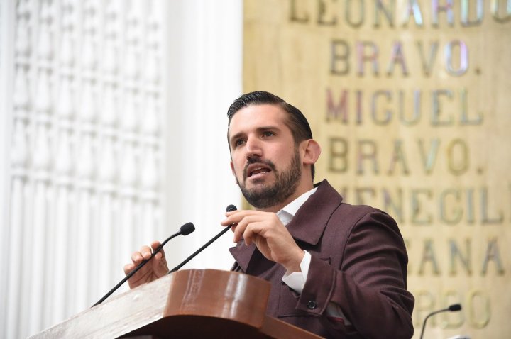 Hombre de pie en un podio, hablando con un micrófono, en un entorno legislativo.