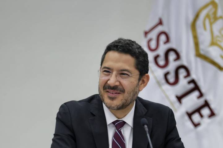 Hombre sonriendo en una reunión, vistiendo un traje oscuro y corbata, con un fondo que incluye el logotipo de ISSSTE.