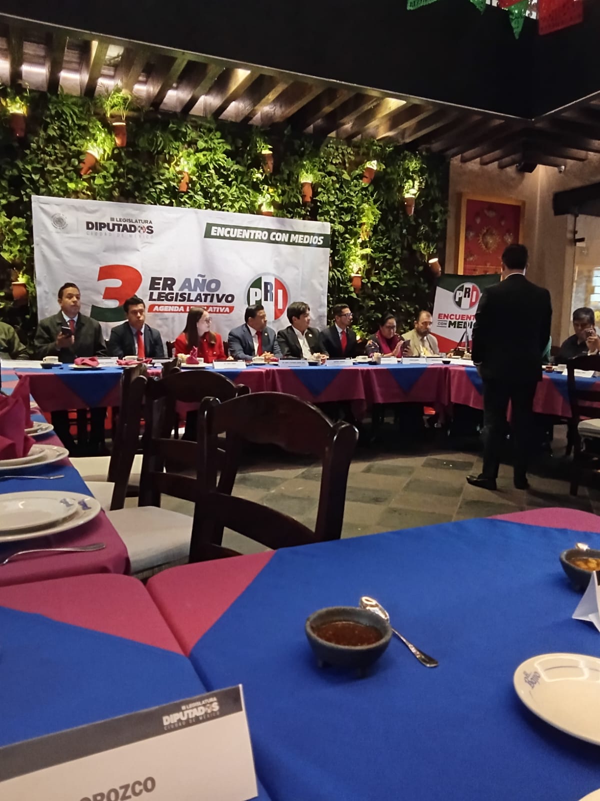 Reunión de diputados en un ambiente formal, con una mesa decorada en colores rosa y azul, y un cartel que indica 'Encuentro con Medios'.