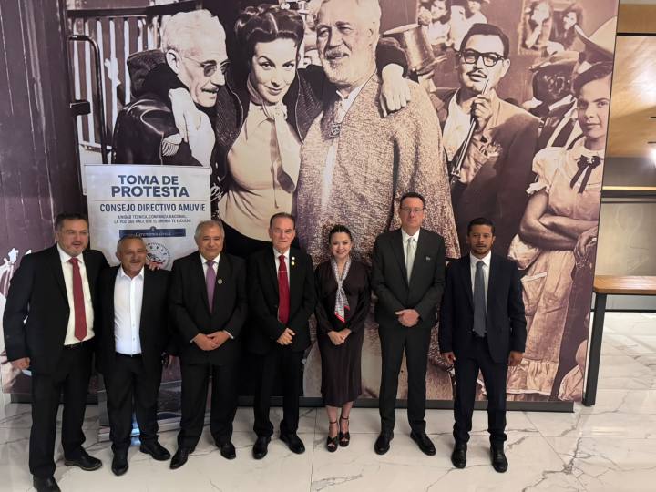 Grupo de personas posando frente a un mural, durante un evento de toma de protesta del Consejo Directivo AMUVIE, con cartel que dice 'TOMA DE PROTESTA'.