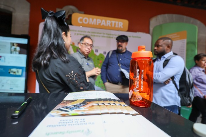 Personas conversando en un evento, con un enfoque en una botella de agua naranja en primer plano y folletos informativos en la mesa.