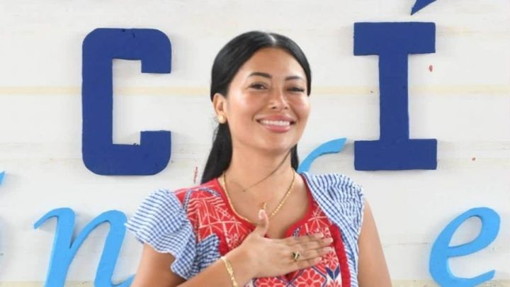 Mujer sonriendo con una mano en el pecho, vestida con una blusa colorida, frente a una pared con letras azules.