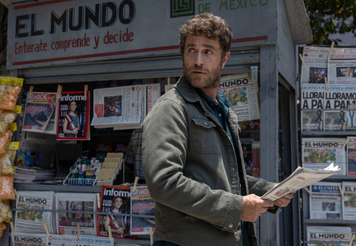 Hombre con chaqueta oscura sosteniendo un periódico frente a un quiosco de noticias con carteles y revistas.
